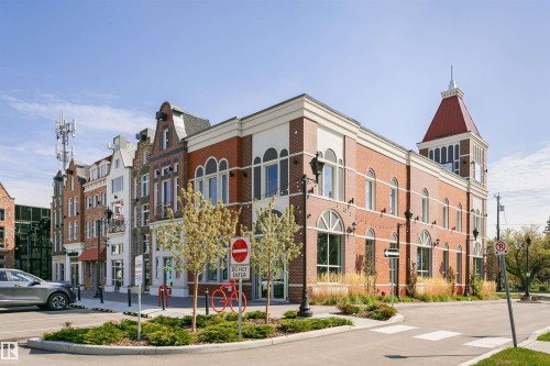 Brick facade building featuring a prominent clock tower with a red roof, arched windows, decorative string lighting, and street-level landscaping - #106 12110 106 Av Nw, Edmonton, AB - Outdoor With Facade