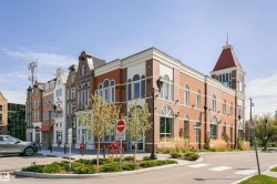Brick facade building featuring a prominent clock tower with a red roof, arched windows, decorative string lighting, and street-level landscaping - 