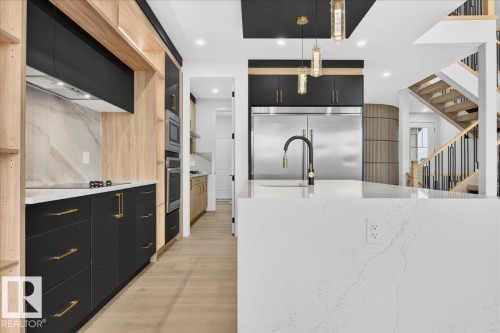 Modern kitchen featuring a large white countertop island, integrated stainless steel refrigerator, black cabinetry with gold-tone hardware, wood-finish flooring, and a contemporary black and gold-tone faucet - 427 172 Avenue Nw, Edmonton, AB - Indoor Photo Showing Kitchen