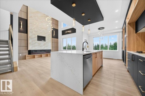 Open-concept living area featuring a kitchen island with integrated dishwasher, light wood-finish flooring, a two-story fireplace with integrated shelving, and large windows - 427 172 Avenue Nw, Edmonton, AB - Indoor Photo Showing Kitchen With Upgraded Kitchen