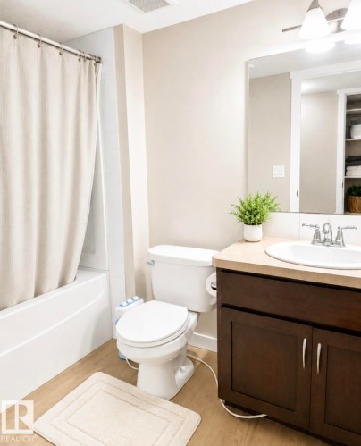 Bathroom featuring wood-finish flooring, a white bathtub with tile surround, a dark wood-finish vanity with a light-toned countertop, a rectangular mirror, and a white toilet - 5135 1A Avenue Sw, Edmonton, AB - Indoor Photo Showing Bathroom