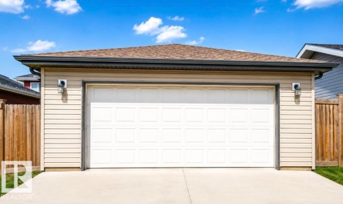 Spacious two-car garage featuring a white sectional overhead door with modern gray trim, flanked by sconce lighting and finished concrete apron - 5135 1A Avenue Sw, Edmonton, AB - Outdoor