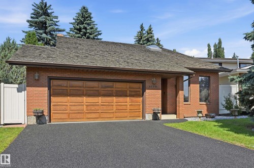 Brick exterior featuring a wood-finish garage door, dark shingle roof, paved driveway, and established landscaping - 4108 Ramsay Road Nw, Edmonton, AB - Outdoor