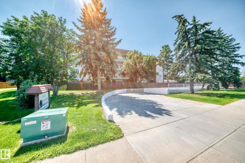 Paved concrete driveway leading to a multi-story building, surrounded by mature trees and a vibrant green lawn - #401 14810 51 Av Nw, Edmonton, AB - Outdoor