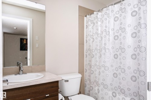 Bathroom featuring a wood-finish vanity with an integrated sink and chrome faucet, a wall-mounted mirror, and a white toilet - #104 9519 160 Av Nw, Edmonton, AB - Indoor Photo Showing Bathroom