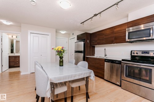 Open-concept kitchen and dining area featuring wood-finish flooring, stainless steel appliances, dark wood-finish cabinetry, light countertops, and track lighting - #104 9519 160 Av Nw, Edmonton, AB - Indoor