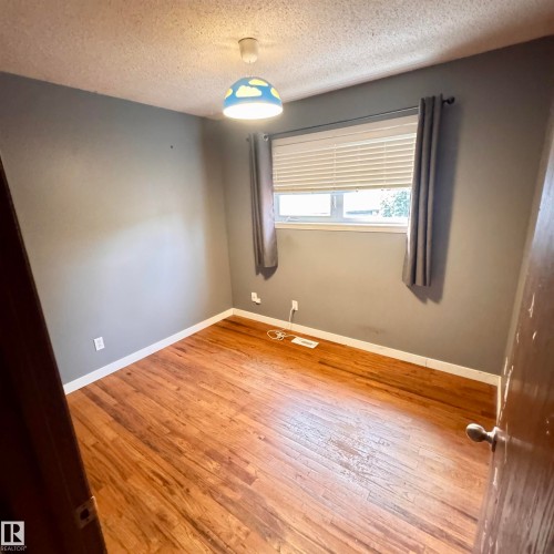 Bright bedroom featuring warm wood-finish flooring, neutral wall paint, and large window with cellular shade and grommet curtain panels - 4821 11A Avenue Nw, Edmonton, AB - Indoor Photo Showing Other Room
