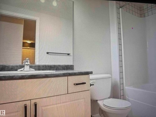 Bathroom featuring a light-toned vanity with dark hardware, a dark countertop, an integrated sink with a chrome faucet, a large wall mirror, and a white toilet - #106 592 Hooke Rd Nw, Edmonton, AB 