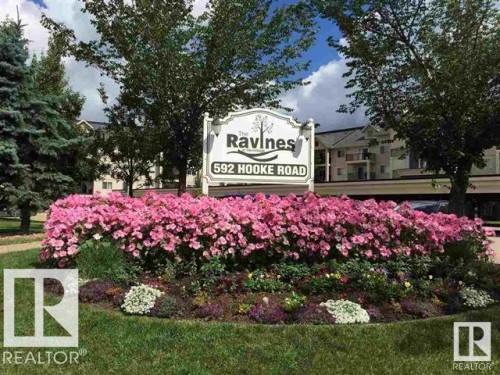 Entry monument with a vibrant floral display featuring pink blooms, evergreen trees, and deciduous trees - #106 592 Hooke Rd Nw, Edmonton, AB 