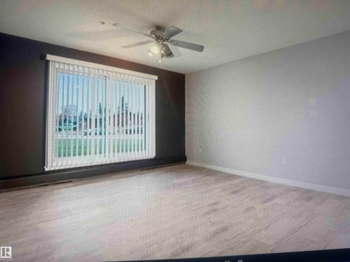Spacious room with wood-finish flooring, a large window with vertical blinds, and a ceiling fan - #106 592 Hooke Rd Nw, Edmonton, AB 