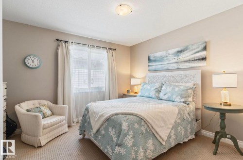 Serene primary bedroom featuring a white-painted headboard, layered bedding in soft blue and neutral tones, dual nightstands with table lamps, and large windows dressed with sheer curtains and drapery panels - 22 Voleta Crest, Spruce Grove, AB - Indoor Photo Showing Bedroom