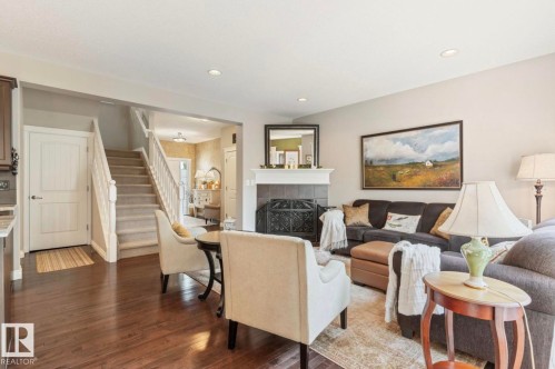 Open-concept living area featuring hardwood flooring, a cream-upholstered accent chair, dark upholstered sofa, wood-burning fireplace with dark surround, and recessed ceiling lighting - 22 Voleta Crest, Spruce Grove, AB - Indoor Photo Showing Living Room With Fireplace