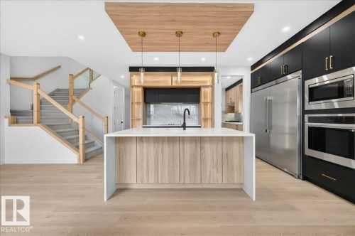 Open-concept kitchen with a large central island, light wood-finish flooring, and a contemporary staircase featuring glass panels and natural wood railings - 403 172 Avenue Nw, Edmonton, AB - Indoor Photo Showing Kitchen With Upgraded Kitchen