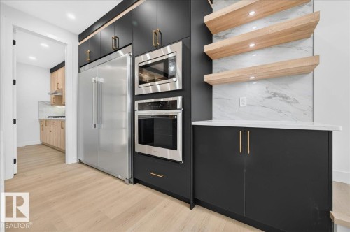Sleek kitchen featuring dark cabinetry with gold-toned hardware, integrated stainless steel appliances, light wood-finish flooring, and floating shelves above a stone-look backsplash - 403 172 Avenue Nw, Edmonton, AB - Indoor