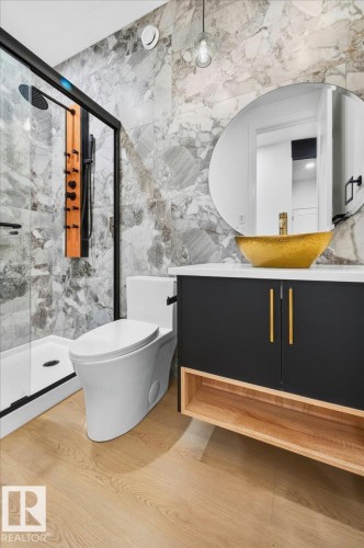Modern bathroom featuring large-format marbled wall tile, light wood-finish flooring, a wall-mounted vanity with black cabinetry and wood-finish shelving, a vessel sink with a gold finish, and a glass-enclosed shower with a multi-function shower panel - 403 172 Avenue Nw, Edmonton, AB - Indoor Photo Showing Bathroom