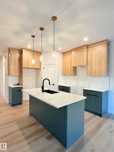 Contemporary kitchen featuring a large island with a white countertop and integrated sink, blonde wood-finish flooring, two-tone cabinetry, a tile backsplash, and pendant lighting - 3150 Magpie Way Nw, Edmonton, AB - Indoor Photo Showing Kitchen
