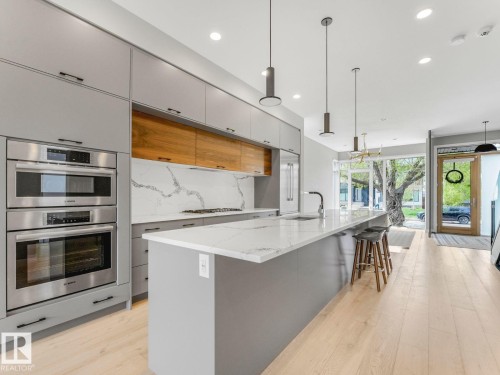 Modern kitchen featuring light gray cabinetry with wood-finish accents, a substantial center island with seating, a gas cooktop, and integrated stainless steel double ovens - 11613 72 Avenue Nw, Edmonton, AB - Indoor Photo Showing Kitchen With Upgraded Kitchen