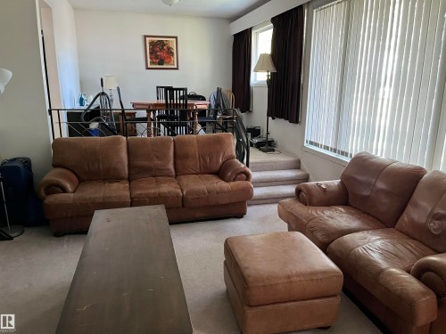 Spacious living area featuring comfortable seating arrangements, neutral carpet flooring, and abundant natural light from expansive windows with vertical blinds - 12011 87 Avenue Nw, Edmonton, AB - Indoor Photo Showing Living Room