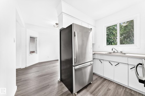 Kitchen area featuring wood-finish flooring, white cabinetry with black hardware, a stainless steel refrigerator, and a sink with a window overhead - 5519 142 Avenue Nw, Edmonton, AB - Indoor Photo Showing Kitchen