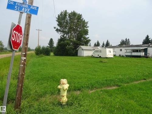 Expansive green space featuring a utility pole with street signs, a yellow fire hydrant, and distant residential structures with mature trees - 5140 51 Avenue, Vilna, AB 
