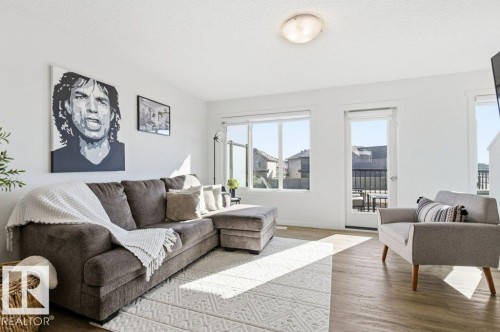 Light wood-finish flooring with white walls and trim, featuring a ceiling-mounted light fixture, a large window, and a glass-paneled door leading to an outdoor area with a black railing - #49 850 Secord Bv Nw, Edmonton, AB - Indoor Photo Showing Living Room