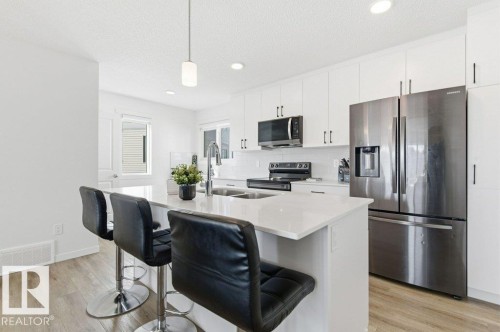 Modern kitchen featuring a central island with countertop seating, stainless steel appliances, white cabinetry with dark hardware, and light wood-finish flooring - #49 850 Secord Bv Nw, Edmonton, AB - Indoor Photo Showing Kitchen With Double Sink