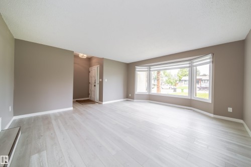 Spacious living area featuring light wood-finish flooring, a large bay window with horizontal blinds, and neutral-toned wall paint - 6907 19 Avenue Nw, Edmonton, AB - Indoor
