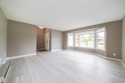 Spacious living area featuring light wood-finish flooring, a large bay window with horizontal blinds, and neutral-toned wall paint - 