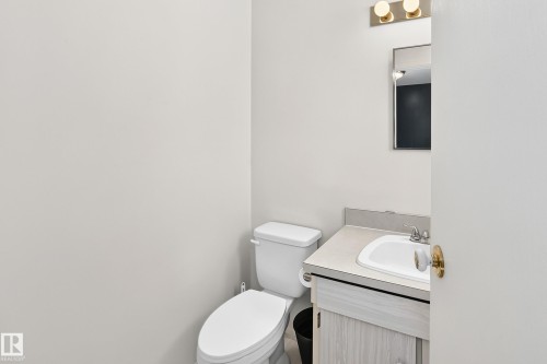 Bathroom featuring a white toilet, light-toned wood-finish vanity, integrated sink, and a wall-mounted mirror with brass-finish lighting - 290 Lancaster Terrace Nw, Edmonton, AB - Indoor Photo Showing Bathroom