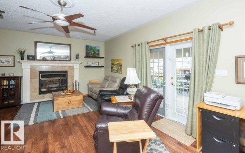 Living area featuring wood-finish flooring, a fireplace with a stone tile surround and white mantel, a ceiling fan, and double glass-paneled doors opening to an outdoor patio - 10711 108 Avenue, Westlock, AB - Indoor Photo Showing Living Room With Fireplace