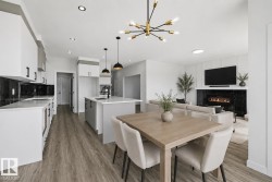 Open-concept living space featuring wood-finish flooring, a contemporary kitchen with white cabinetry and dark backsplash, a kitchen island with sink and pendant lighting, a dining area, and a living area with a fireplace - 