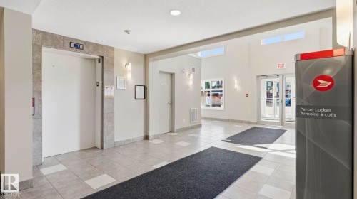 Spacious building lobby featuring light-colored floor tiling, a modern elevator with a stone-tile surround, ample natural light from upper windows, and a parcel locker - #361 7805 71 St Nw, Edmonton, AB - Indoor Photo Showing Other Room