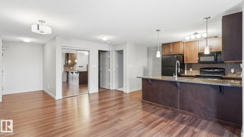 Open-concept living space featuring wood-finish flooring, a kitchen island with stone countertop, stainless steel sink with gooseneck faucet, black appliances, and pendant lighting - #361 7805 71 St Nw, Edmonton, AB - Indoor Photo Showing Kitchen