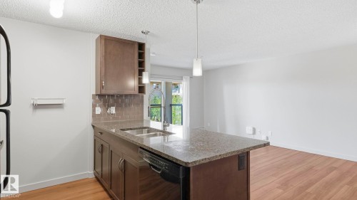Modern kitchen featuring a substantial island with granite countertop and dark cabinetry, stainless steel appliances, pendant lighting, and abundant cabinet storage - #361 7805 71 St Nw, Edmonton, AB - Indoor Photo Showing Kitchen With Double Sink
