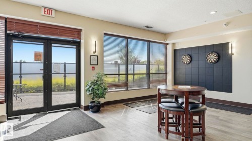 Lobby area featuring wood-finish flooring, large windows with horizontal blinds, double glass entry doors, and a decorative wall panel with integrated lighting - #361 7805 71 St Nw, Edmonton, AB - Indoor Photo Showing Other Room