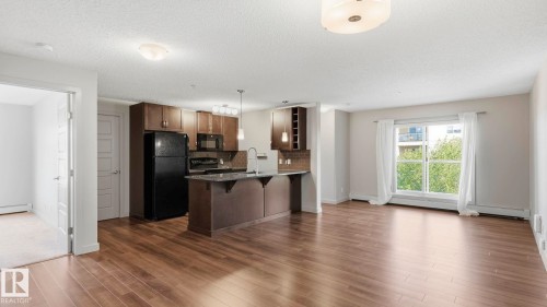 Open-concept kitchen and living area featuring dark cabinetry with stainless steel appliances, a center island with breakfast bar, wood-finish flooring throughout, recessed ceiling lighting, and abundant natural light from multiple windows and glass doors - #361 7805 71 St Nw, Edmonton, AB - Indoor Photo Showing Kitchen