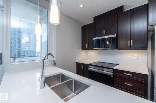 Modern kitchen featuring dark wood cabinetry, stainless steel appliances, a white subway tile backsplash, and white countertops with an undermount double-basin sink - #1201 10238 103 St Nw, Edmonton, AB - Indoor Photo Showing Kitchen With Double Sink