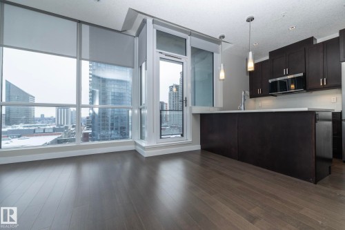 Open-concept living area featuring extensive window walls, wood-finish flooring, a kitchen island with pendant lighting, dark wood cabinetry, and stainless steel appliances - #1201 10238 103 St Nw, Edmonton, AB - Indoor Photo Showing Kitchen With Upgraded Kitchen