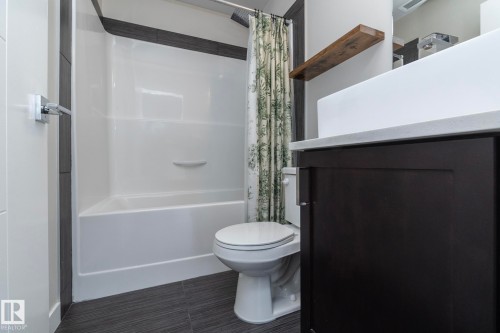 Bathroom featuring a white bathtub/shower combination with dark accent tile, a white toilet, a dark wood-finish vanity with a light countertop, and a wood-finish floating shelf - #1201 10238 103 St Nw, Edmonton, AB - Indoor Photo Showing Bathroom
