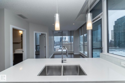 Large kitchen island with a double stainless steel sink, polished chrome faucet, white countertop, and two pendant lights - #1201 10238 103 St Nw, Edmonton, AB - Indoor Photo Showing Kitchen With Double Sink