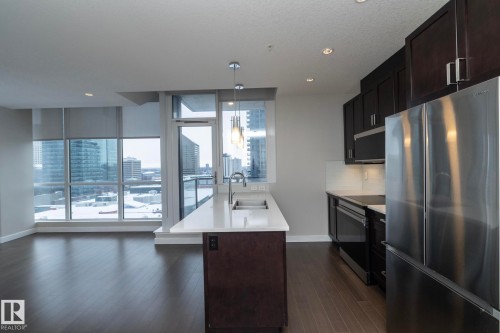 Open-concept living area featuring wood-finish flooring, a contemporary kitchen island with seating, pendant lighting, stainless steel appliances, and extensive windows with roller shades - #1201 10238 103 St Nw, Edmonton, AB - Indoor Photo Showing Kitchen With Stainless Steel Kitchen With Double Sink With Upgraded Kitchen