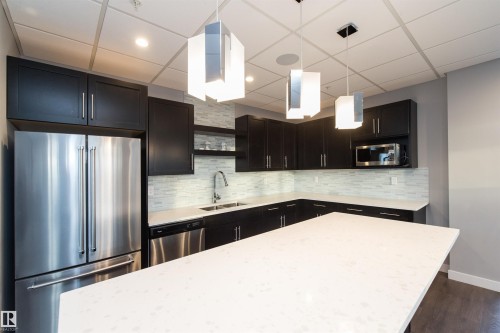 Contemporary kitchen featuring dark cabinetry, stainless steel appliances, light-toned countertops, a full-height tile backsplash, and three suspended light fixtures - #1201 10238 103 St Nw, Edmonton, AB - Indoor Photo Showing Kitchen With Stainless Steel Kitchen With Upgraded Kitchen