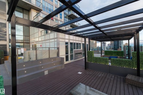 Rooftop deck featuring a black metal pergola, composite wood-finish decking, integrated planter boxes with greenery, a built-in fireplace, and a tiered spa with accent lighting - #1201 10238 103 St Nw, Edmonton, AB - Outdoor