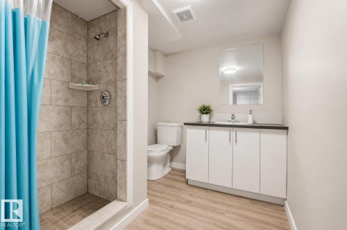 Bathroom featuring a tile-surround shower with a corner shelf, a white vanity with a dark countertop and integrated sink, a wall-mounted mirror, and wood-finish flooring - 12235 104 Street Nw, Edmonton, AB - Indoor Photo Showing Bathroom