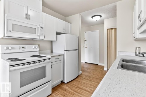 Kitchen featuring white cabinetry, white appliances, wood-finish flooring, and light-colored countertops with an integrated double basin stainless steel sink - #5 11265 31 Av Nw, Edmonton, AB - Indoor Photo Showing Kitchen With Double Sink