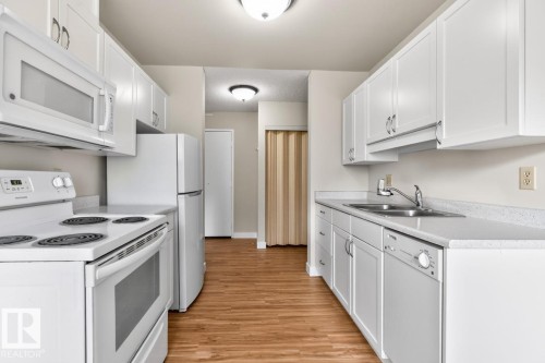 Galley kitchen featuring white cabinetry, wood-finish flooring, white appliances, and a double basin sink - #5 11265 31 Av Nw, Edmonton, AB - Indoor Photo Showing Kitchen With Double Sink