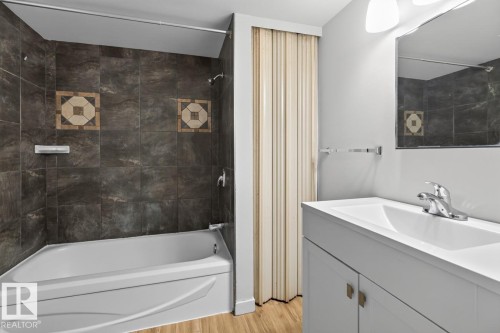 Bathroom featuring a white bathtub, dark tiled shower surround with decorative accents, light wood-finish flooring, and a white vanity with integrated sink and chrome faucet - #5 11265 31 Av Nw, Edmonton, AB - Indoor Photo Showing Bathroom