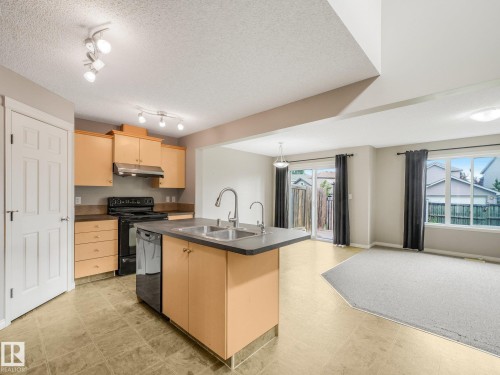 Open-concept kitchen and living area featuring natural wood cabinetry, stainless steel appliances, a dual-basin island with undermount sink, tile countertops, and polished concrete flooring - 11844 21 Avenue Sw, Edmonton, AB - Indoor Photo Showing Kitchen With Double Sink