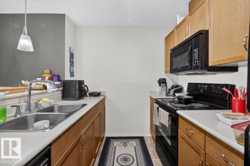 Kitchen featuring wood-finish cabinetry, light-toned countertops, and a full suite of black appliances, including a built-in microwave and electric range - #421 1180 Hyndman Rd Nw, Edmonton, AB - Indoor Photo Showing Kitchen With Double Sink