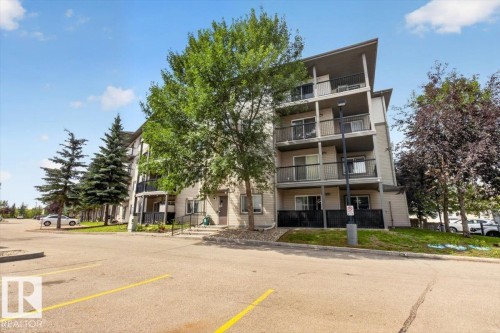 Multi-level building with individual balconies, exterior lighting, and a paved parking area with marked spaces - #421 1180 Hyndman Rd Nw, Edmonton, AB - Outdoor With Facade